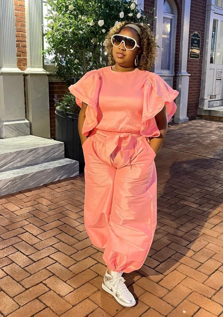 Woman wearing a pink ruffle-sleeve two-piece set with drawstring pants, white and silver sneakers, and oversized sunglasses.
