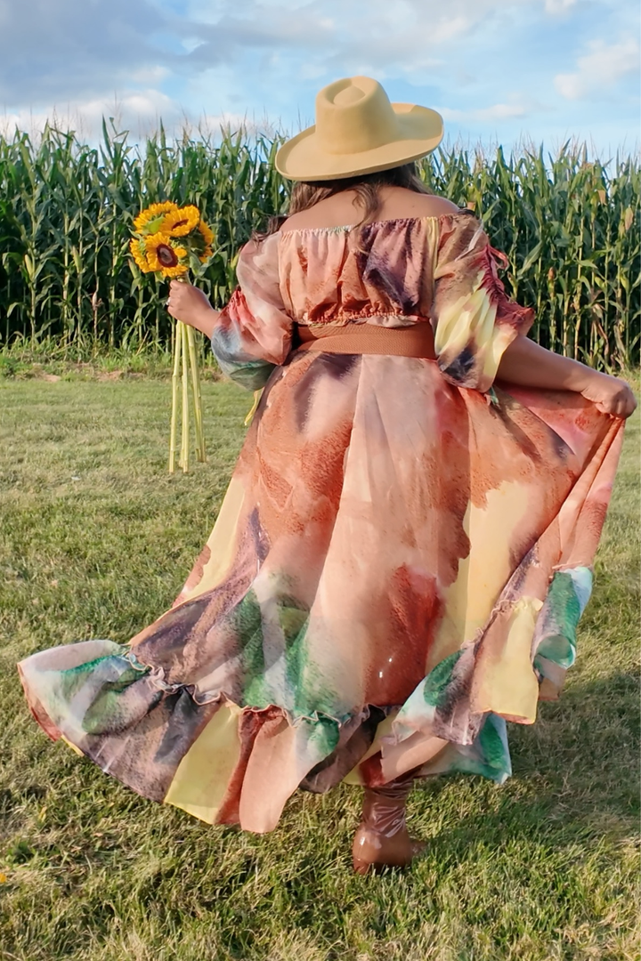 Person in a colorful dress and hat holding sunflowers in a cornfield