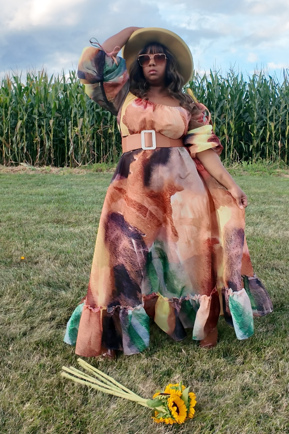 Woman in a colorful dress and hat standing in a field with corn stalks.