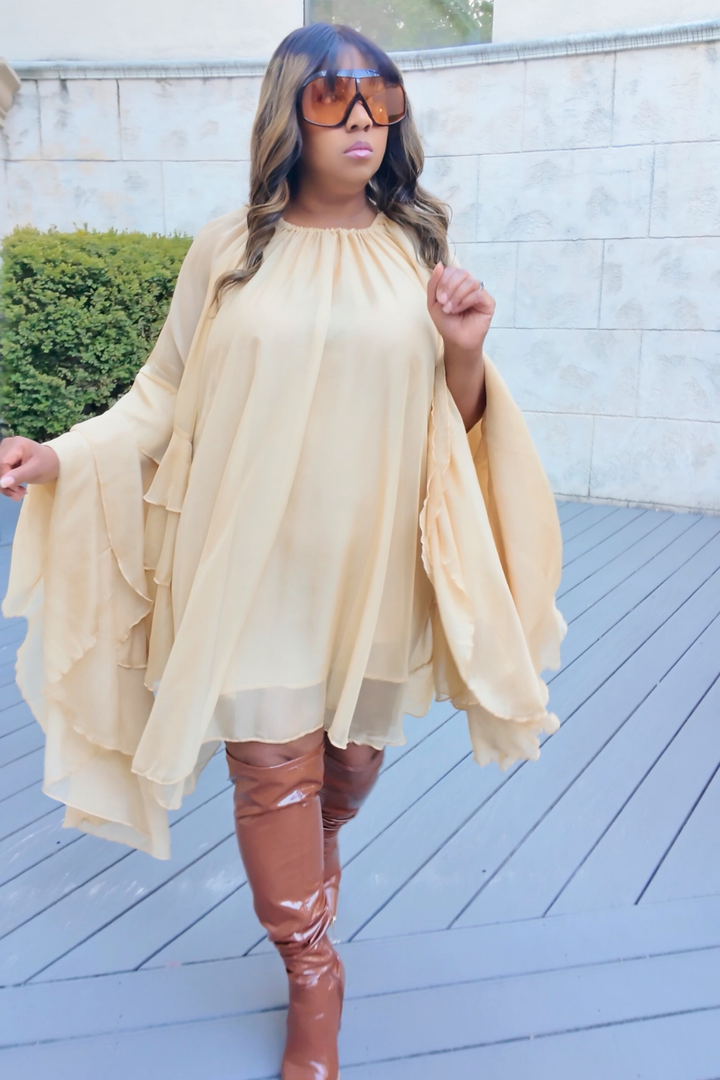 A woman wearing the Golden Cascade Dress in soft champagne chiffon with dramatic layered sleeves, styled with glossy thigh-high boots and oversized gradient sunglasses, posing on a sunlit terrace