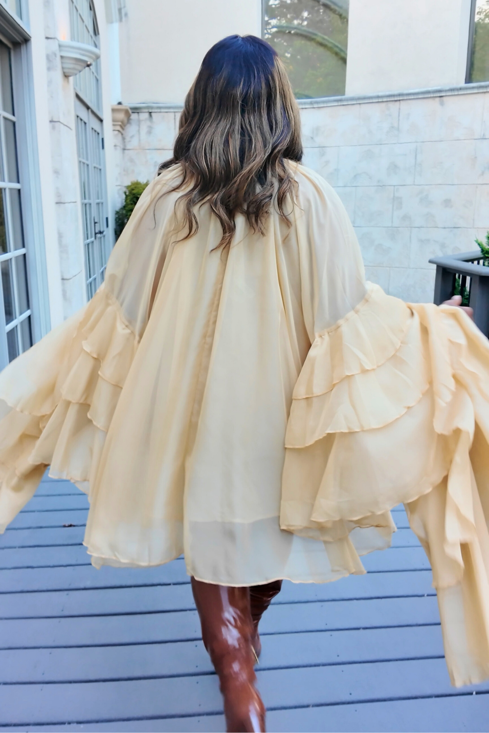 A woman wearing the Golden Cascade Dress in soft champagne chiffon with dramatic layered sleeves, styled with glossy thigh-high boots and oversized gradient sunglasses, posing on a sunlit terrace