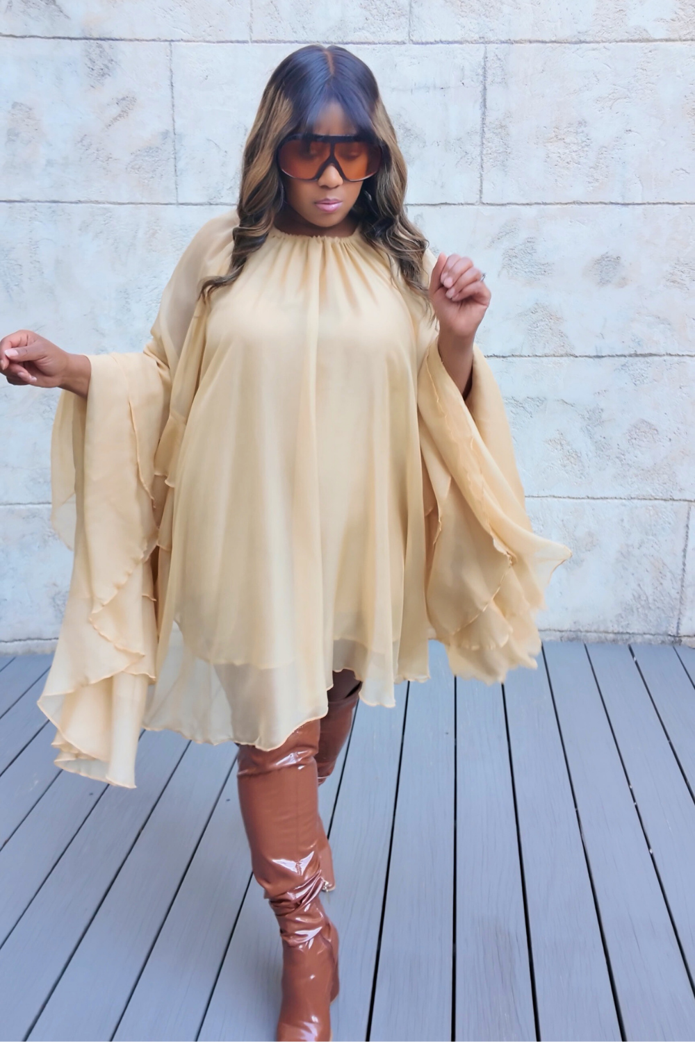 A woman wearing the Golden Cascade Dress in soft champagne chiffon with dramatic layered sleeves, styled with glossy thigh-high boots and oversized gradient sunglasses, posing on a sunlit terrace