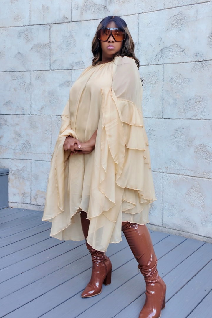 A woman wearing the Golden Cascade Dress in soft champagne chiffon with dramatic layered sleeves, styled with glossy thigh-high boots and oversized gradient sunglasses, posing on a sunlit terrace