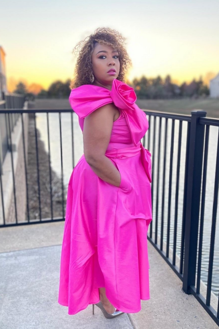 Woman in a bright pink dress with a large bow standing on a balcony at sunset.