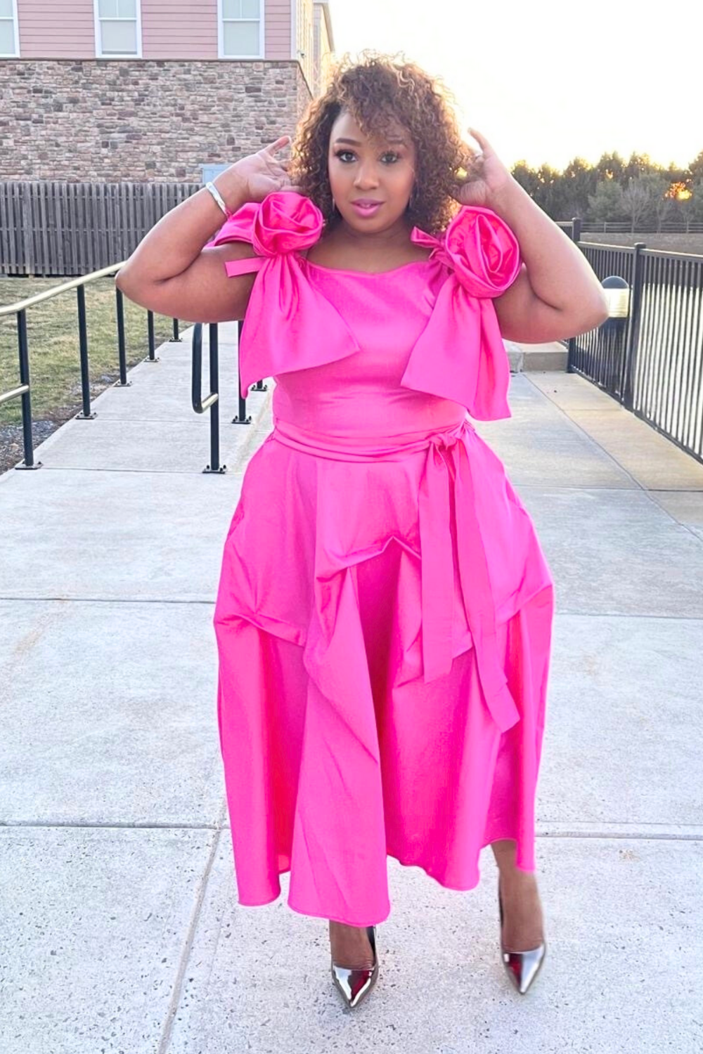 Woman wearing a bright pink dress with ruffled sleeves and a belt, standing on a sidewalk.