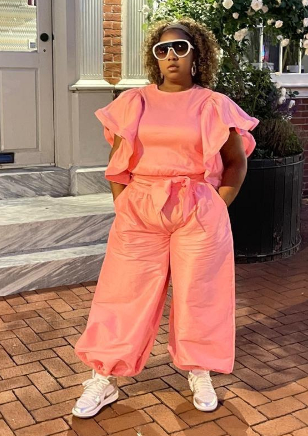 Woman wearing a pink ruffle-sleeve two-piece set with drawstring pants, white and silver sneakers, and oversized sunglasses.