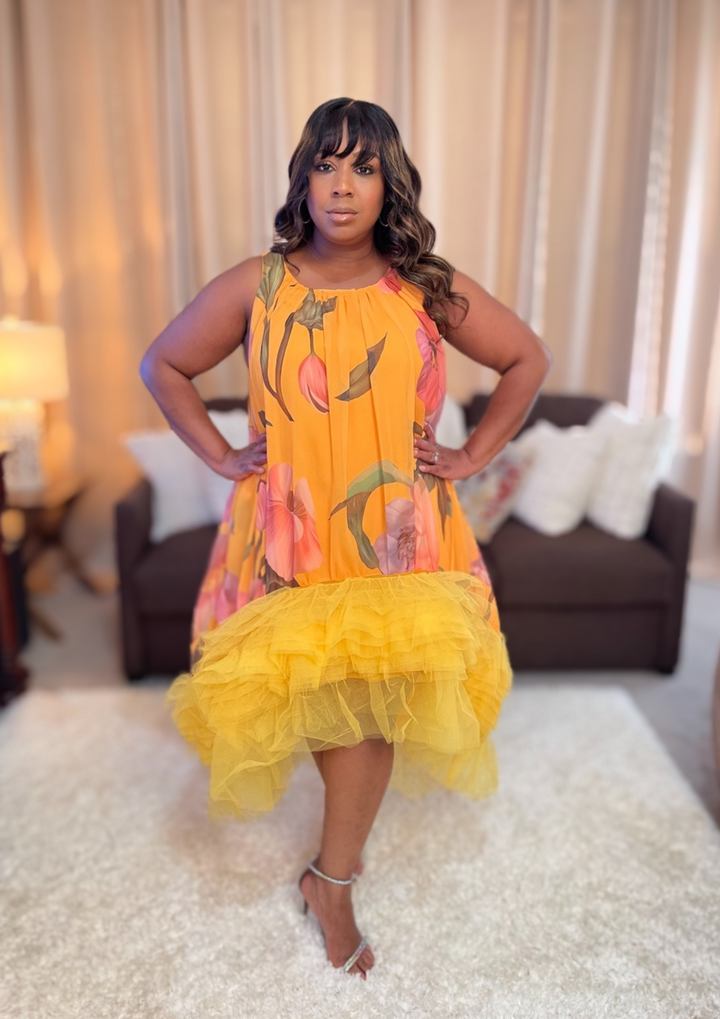 Woman wearing a colorful dress with floral patterns in a room with curtains and furniture.
