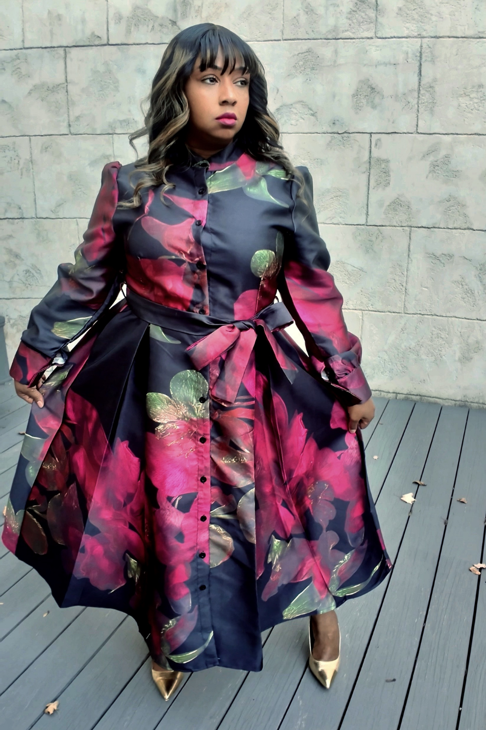 A woman wearing a colorful floral dress standing on a wooden deck with a stone wall background
