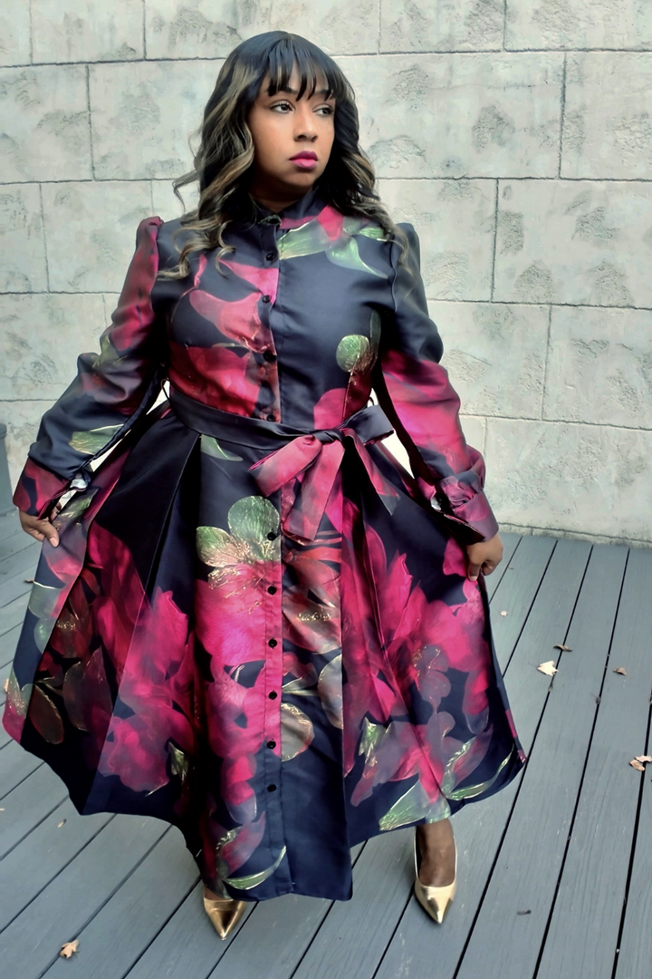 A woman wearing a colorful floral dress standing on a wooden deck with a stone wall background