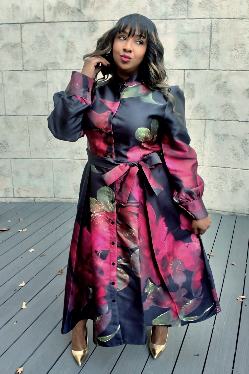 A woman wearing a colorful floral dress standing on a wooden deck with a stone wall background