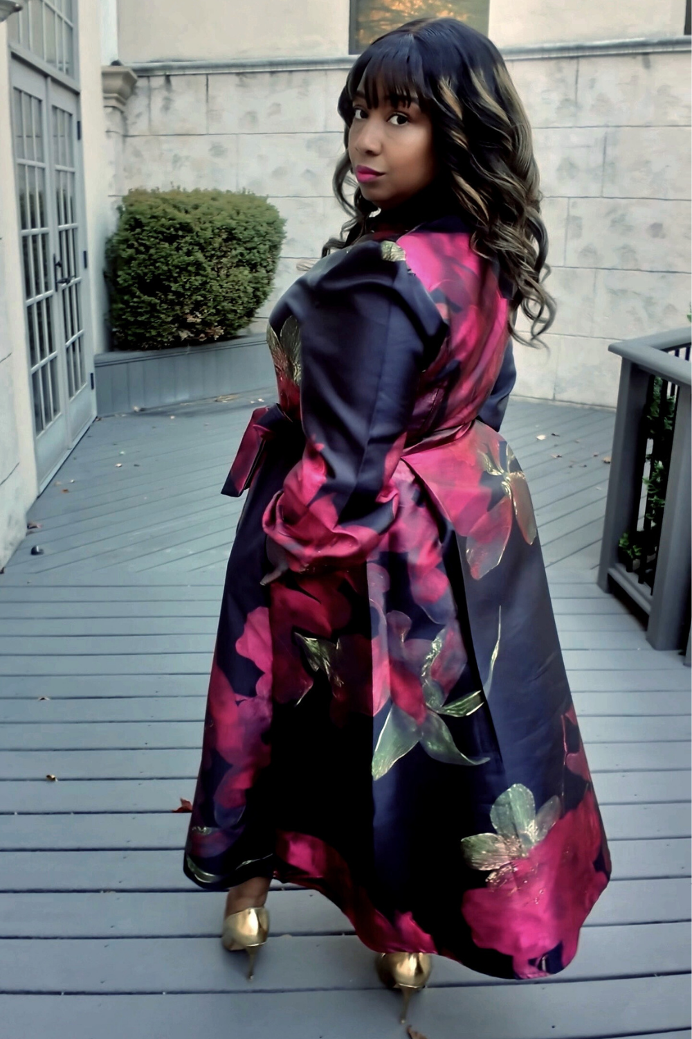 A woman wearing a colorful floral dress standing on a wooden deck with a stone wall background.